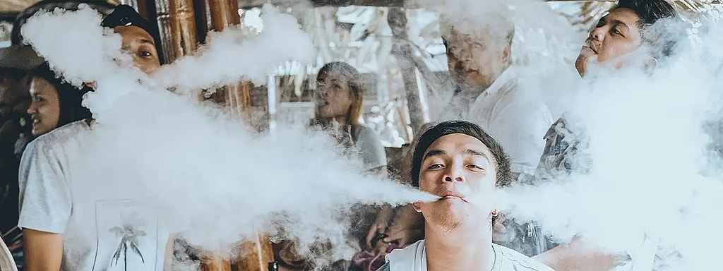 Eine Gruppe von Menschen in entspannter Atmosphäre. Eine Person im Vordergrund atmet Dampf aus einer E-Zigarette aus und erzeugt eine Rauchwolke. Andere stehen in der Nähe, einige blicken in die Kamera, andere stehen abseits.